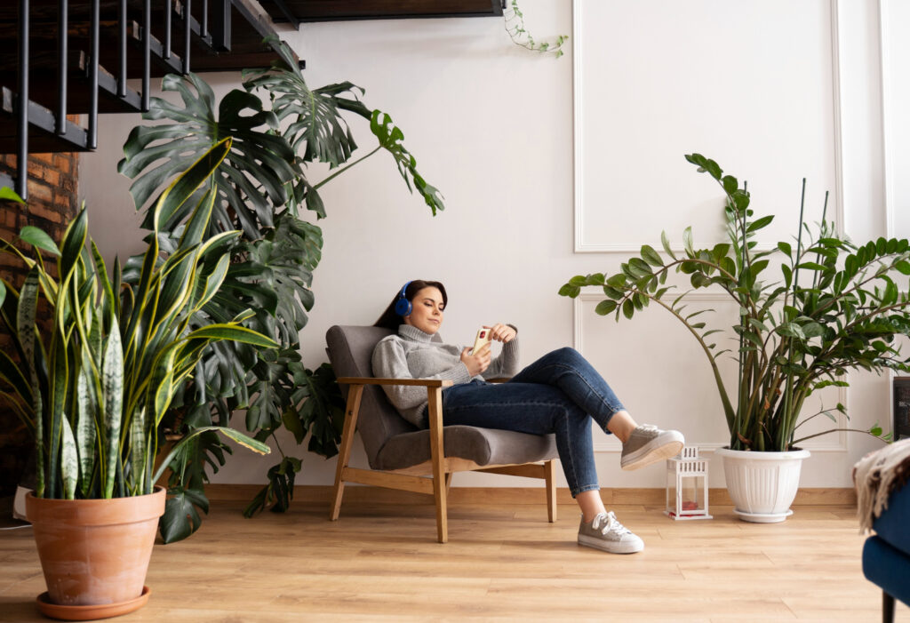 A person sits comfortably in a stylish armchair surrounded by lush indoor plants, enjoying their phone in a cozy room.