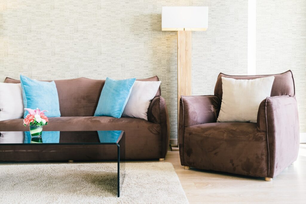 Cozy living room featuring a brown sofa with blue and white pillows, an armchair, a glass coffee table, and a decorative flower arrangement.