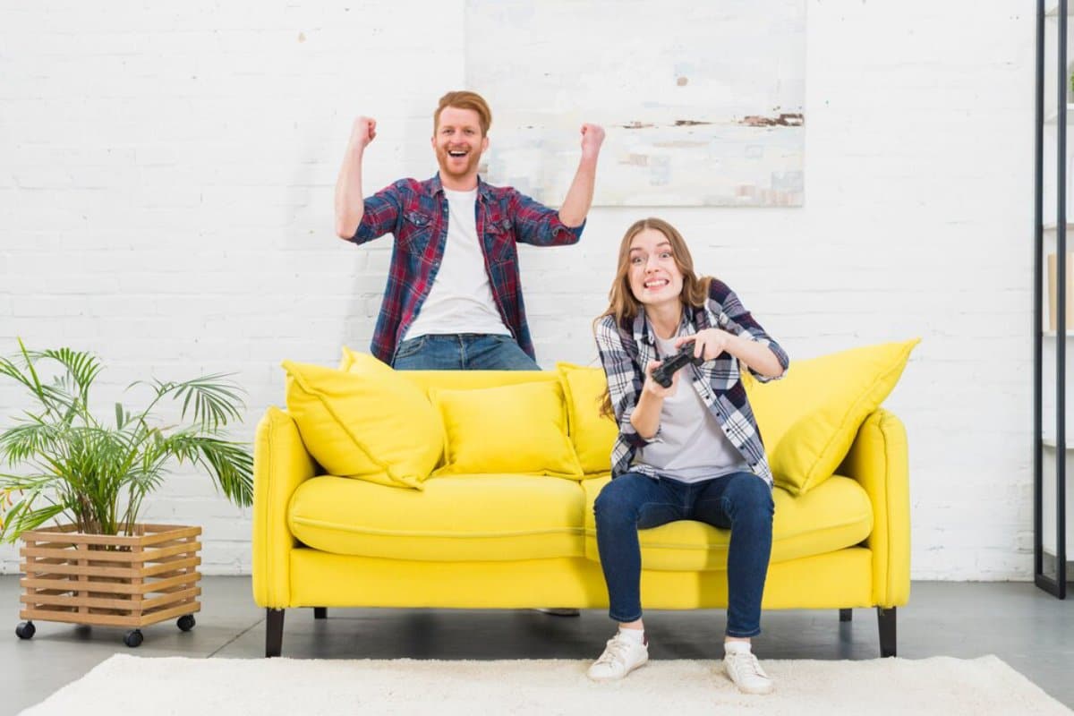 A person joyfully plays a video game on a yellow couch while another cheers enthusiastically in the background.