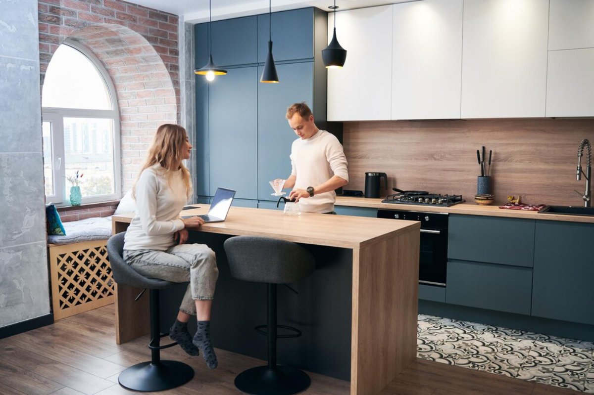 A stylish modern kitchen with a wooden island, pendant lights, and a woman using a laptop while a man prepares a drink nearby.
