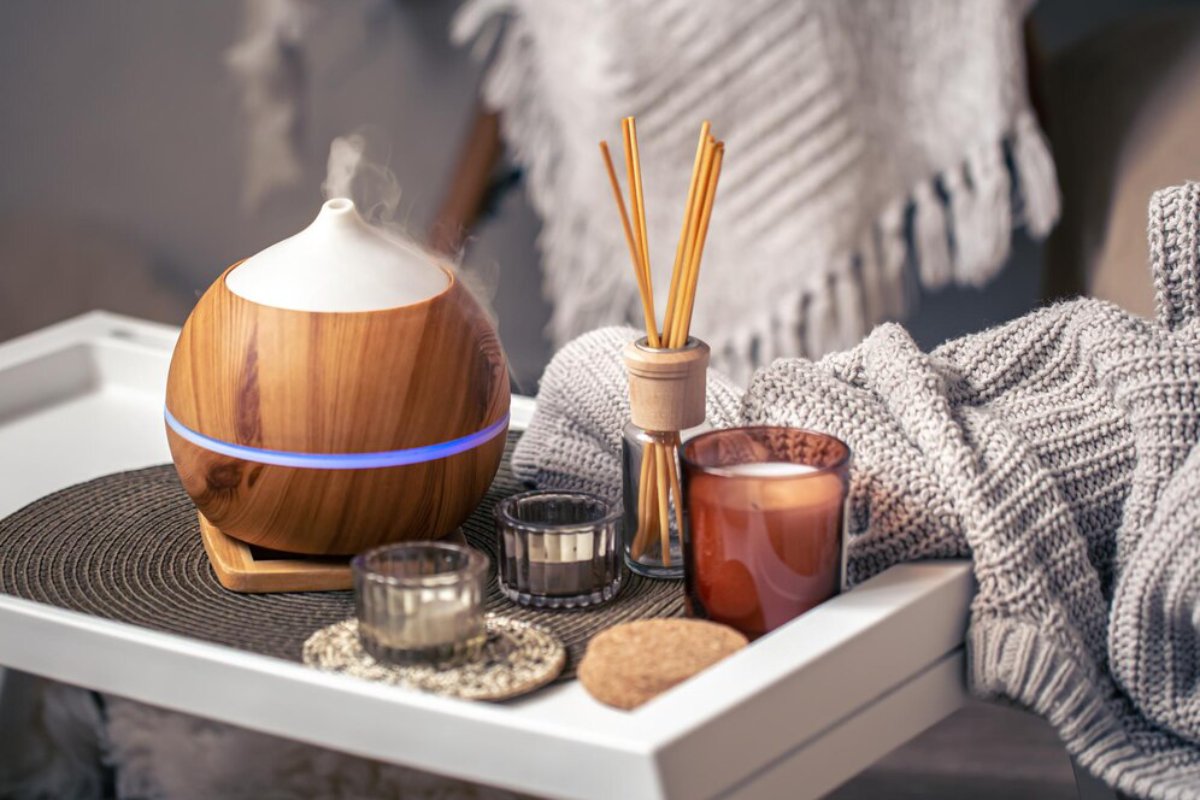 A wooden essential oil diffuser emits steam, surrounded by candles, a blanket, and incense sticks on a cozy tray.