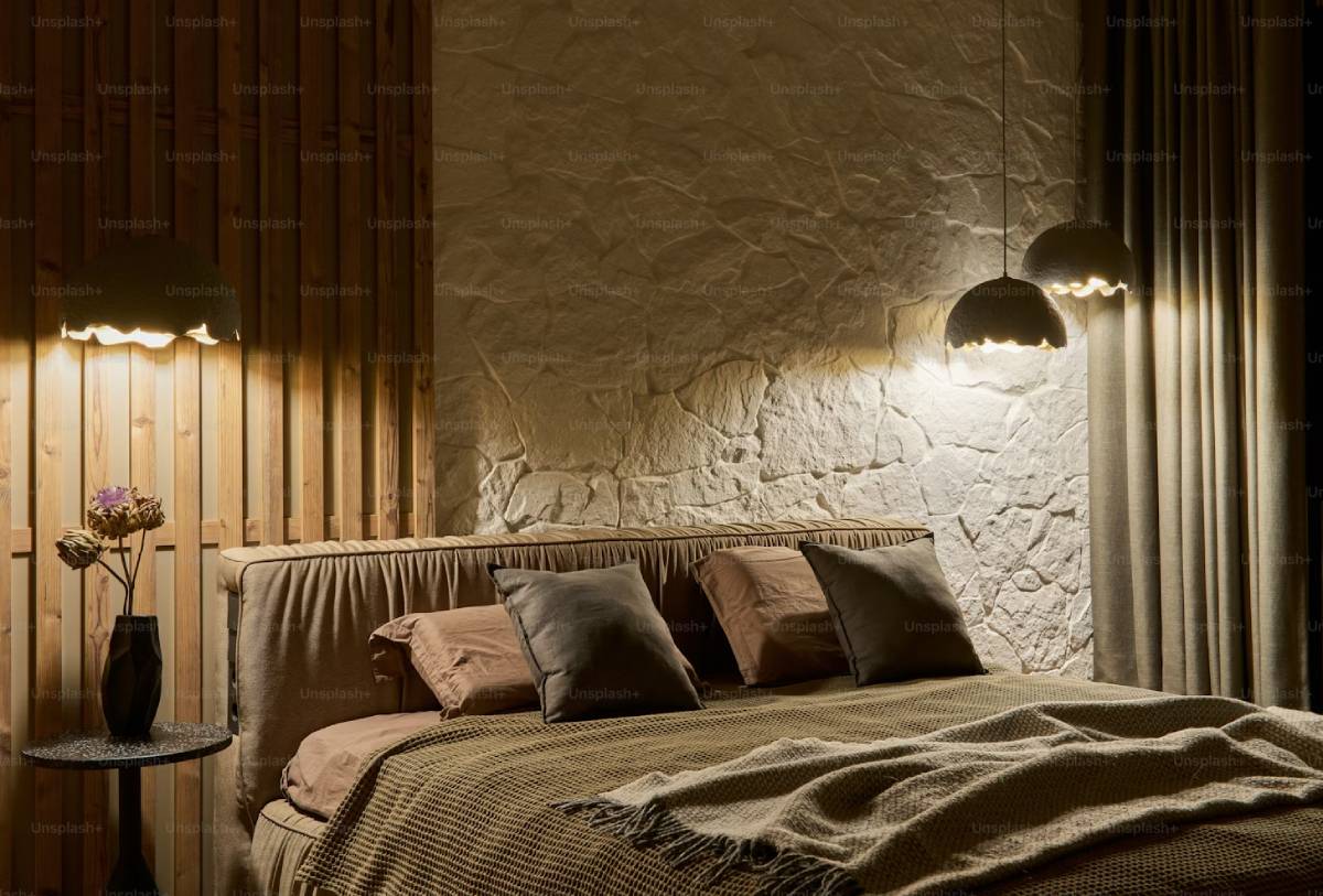 Cozy bedroom with a textured stone wall, soft bedding, decorative pillows, a bedside table with a vase, and ambient pendant lighting.
