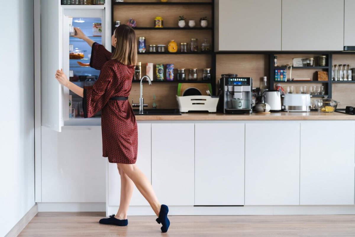 A woman in a red polka dot robe reaches into a refrigerator in a modern kitchen, with shelves containing various food items and appliances.
