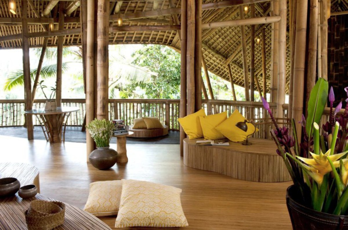 A spacious bamboo interior with wooden floors, bright yellow cushions, and plants, surrounded by large windows and tropical greenery.