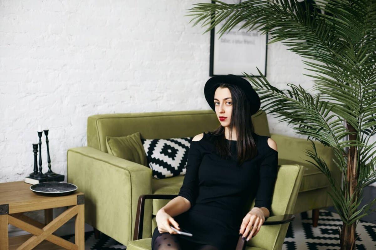 A woman in a black dress and hat sits casually in an olive green chair, surrounded by modern decor and a potted plant.