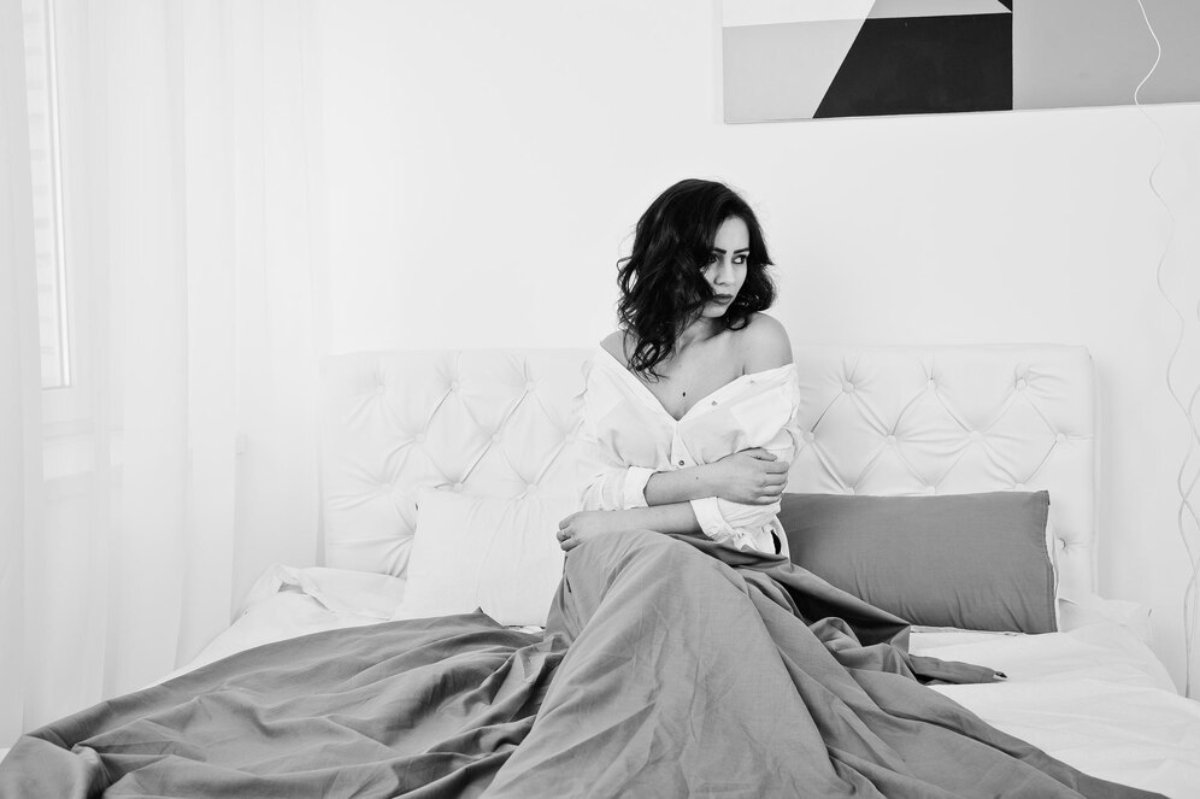 A woman sits on a white tufted bed, draped in a gray blanket, wearing an unbuttoned white shirt, surrounded by soft, sheer curtains.