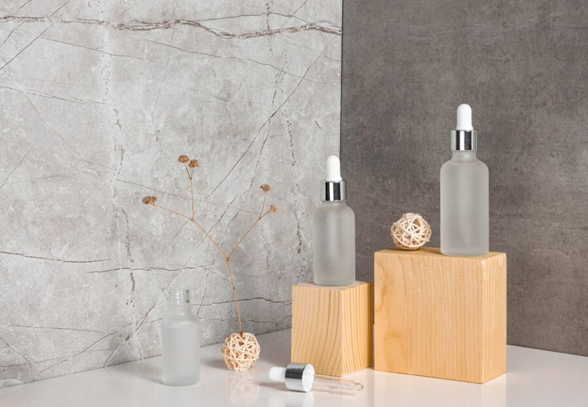 Three frosted glass dropper bottles are displayed on wooden cubes, alongside dried flowers and decorative spheres, against a textured gray backdrop.
