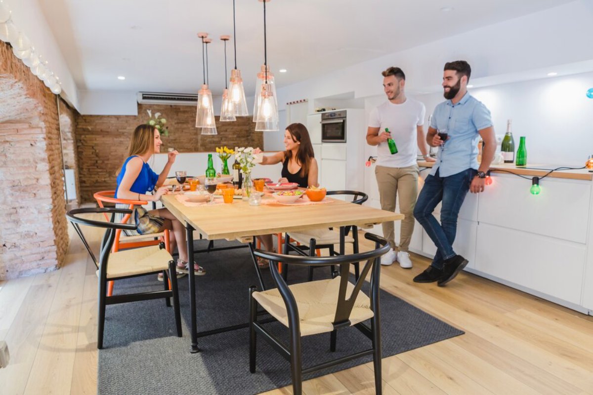 A modern kitchen and dining area with friends enjoying drinks and a meal around a wooden table decorated with flowers and colorful tableware.