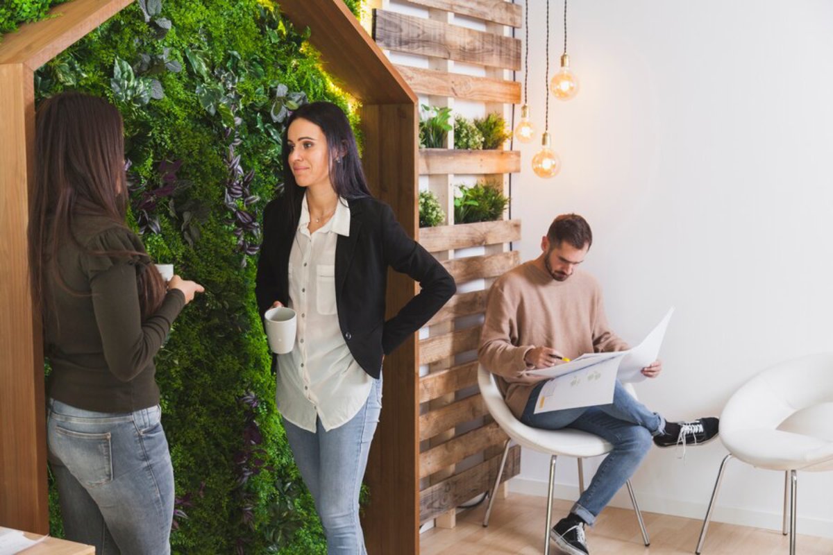 Two people stand engaged in conversation in a modern office with a vertical garden backdrop, while another sits drawing on a notepad.
