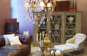 A cozy interior with a brown leather sofa, elegant chandelier, vintage display cabinet, wooden decor, and a light-colored armchair.