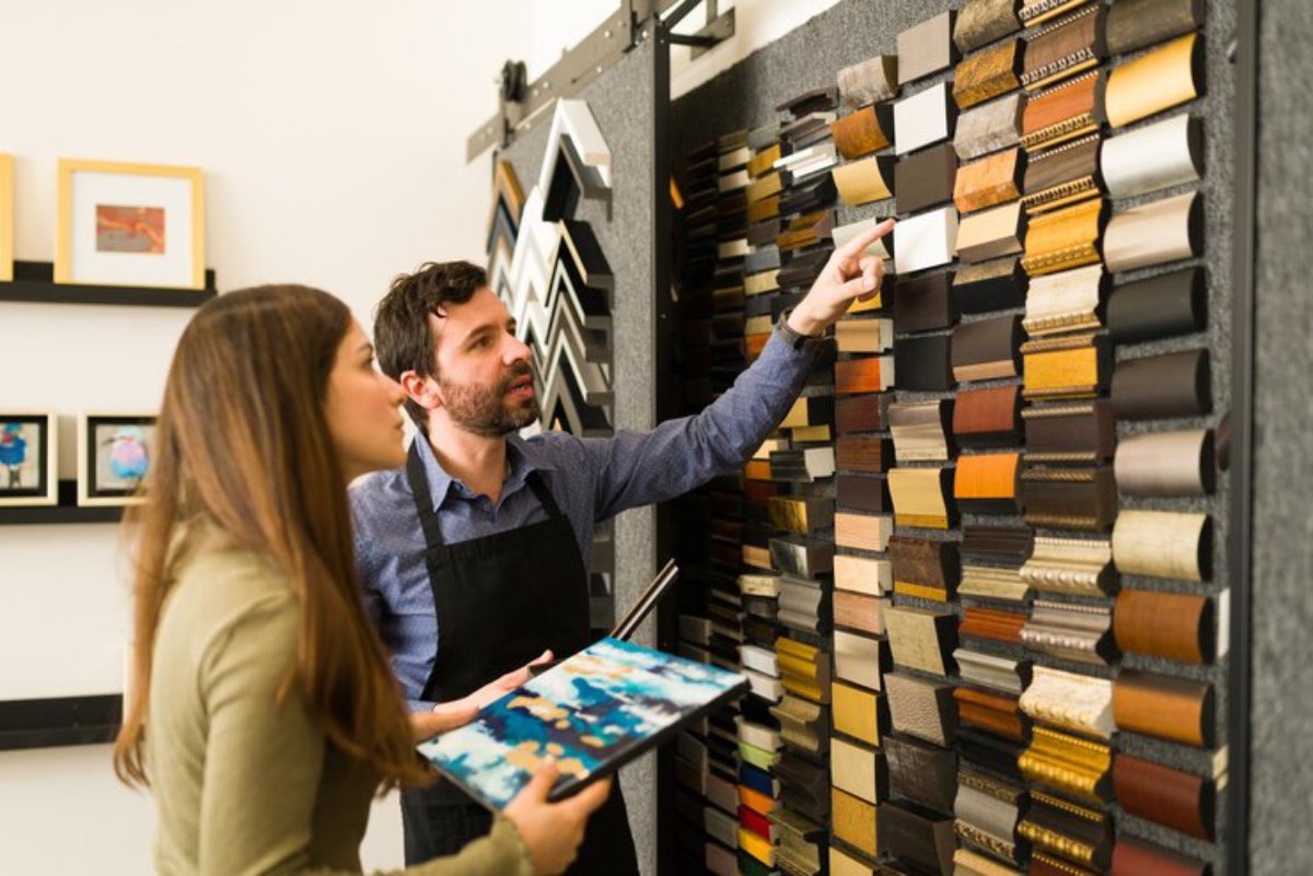 A person examines a colorful selection of picture frame samples with a colleague in a framing shop, discussing design options.