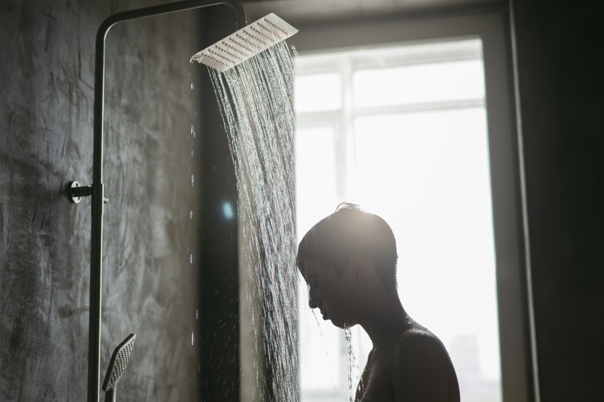 A person stands showering under a modern rain showerhead, water cascading down against a backdrop of a large window.