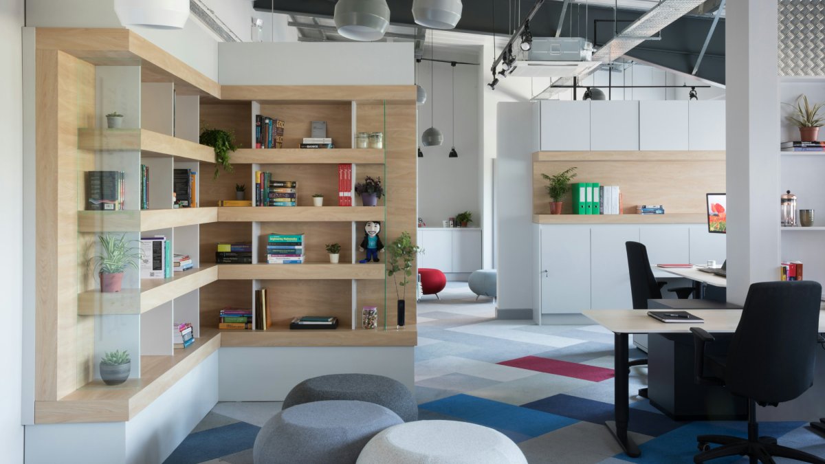 Modern office interior featuring wooden bookshelves, plants, and a desk area with colorful rugs and seating. Bright and inviting atmosphere.
