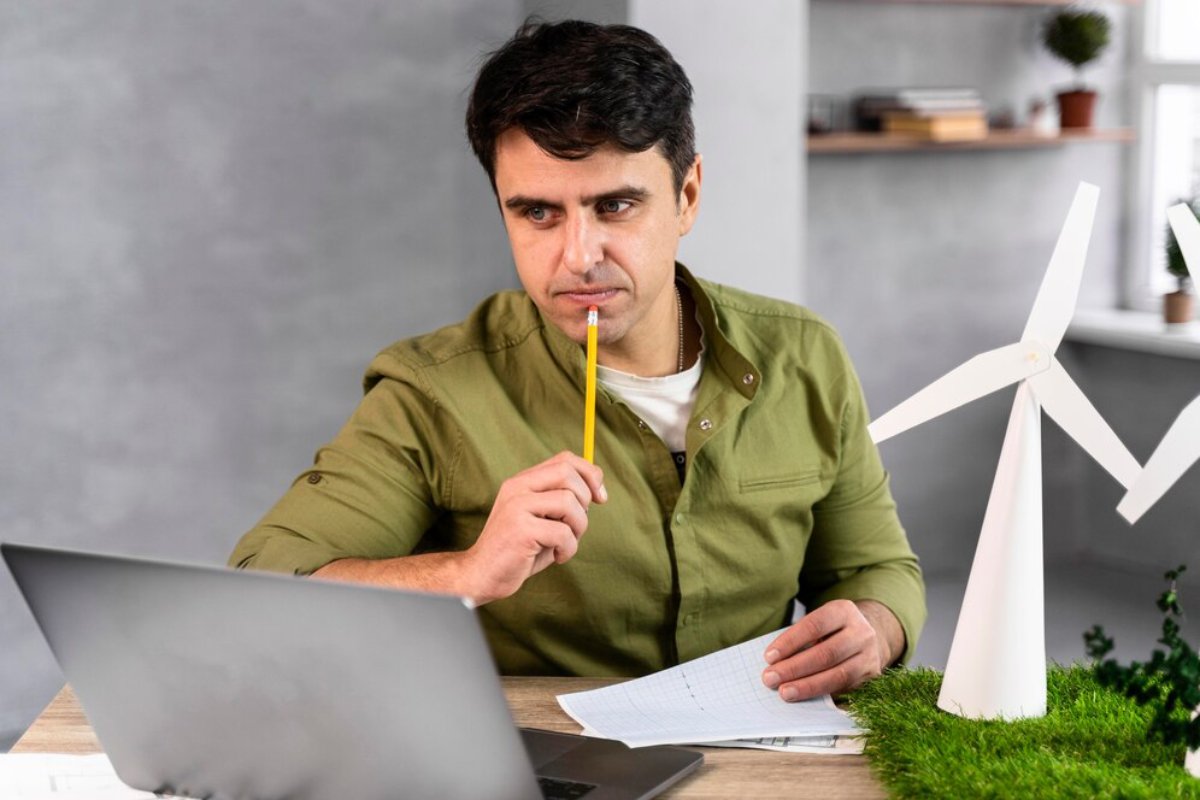 A person in a green shirt is holding a yellow pencil, seated at a desk with a laptop and a small wind turbine model.