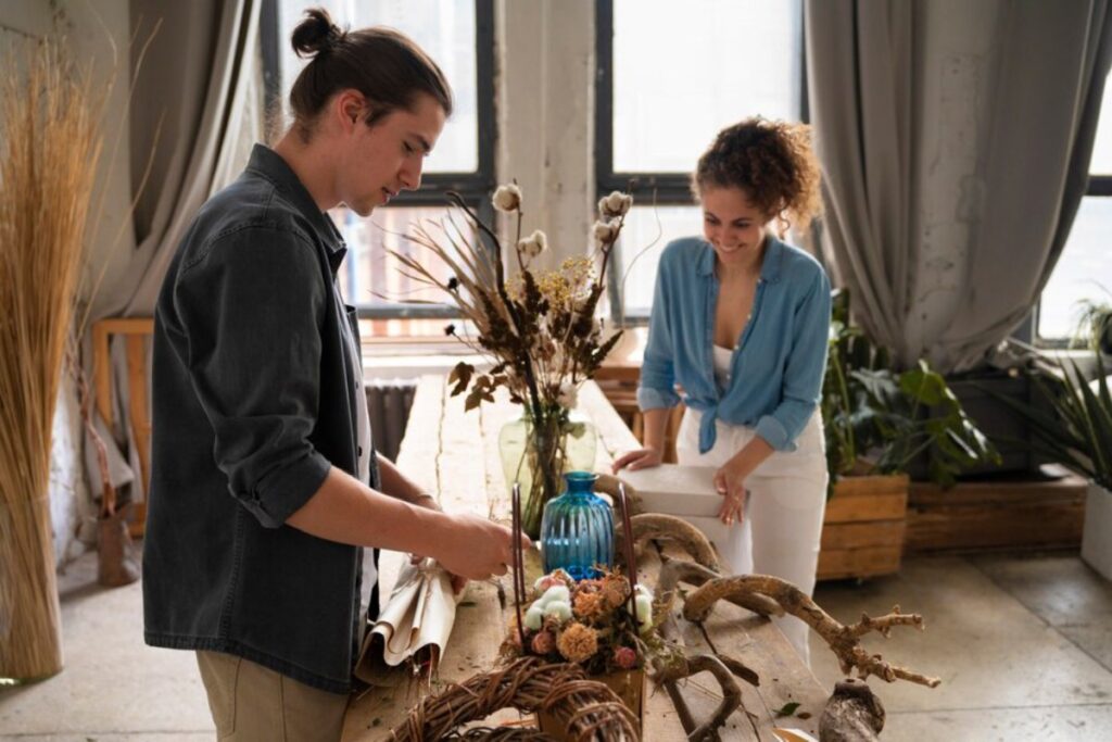 Two individuals are arranging dried flowers and natural materials on a rustic table in a sunlit, stylish indoor space.