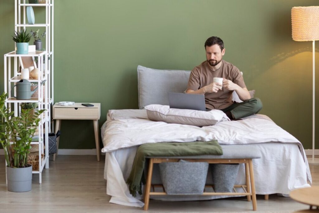 A cozy bedroom scene with a person sitting on a bed, working on a laptop, and holding a cup, surrounded by plants and decorative items.