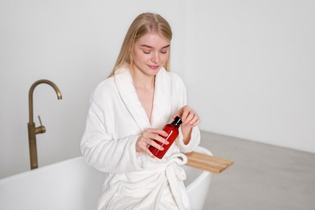 A person in a white robe holds a red bottle while seated by a modern bathtub in a bright, minimalistic bathroom.