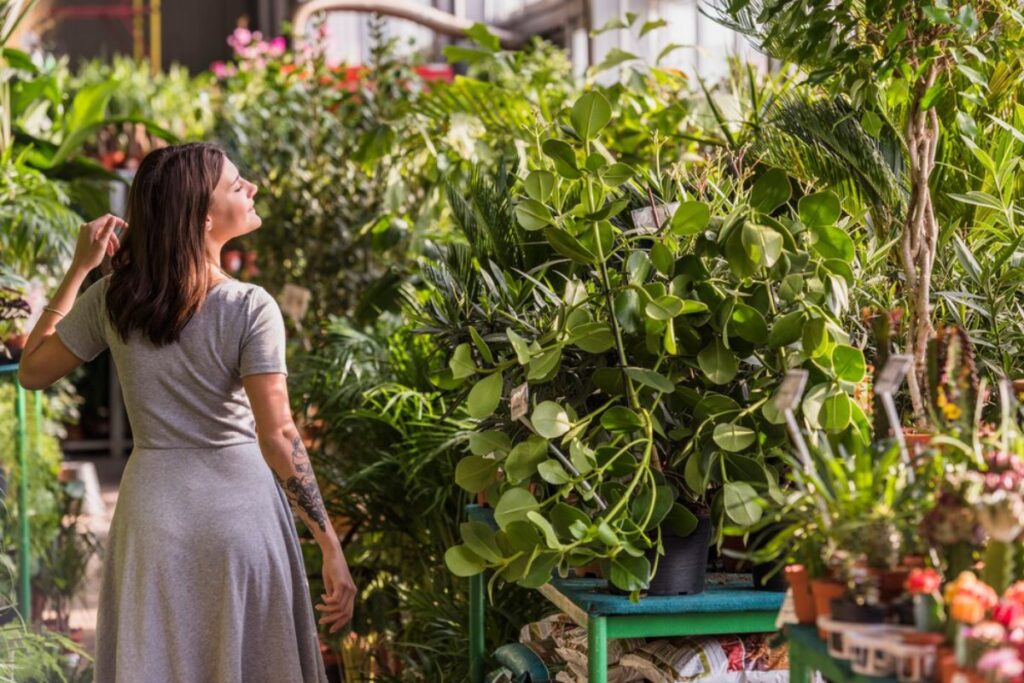 A person in a gray dress gestures thoughtfully while surrounded by vibrant greenery and potted plants in a sunlit greenhouse.