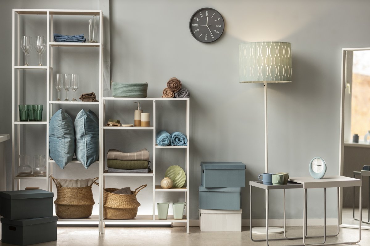 A cozy living space features a white shelving unit filled with decorative boxes, pillows, and glassware, alongside a stylish lamp and clock.