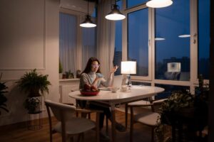 A woman sits at a dining table with a laptop, phone, and a bowl of fruit, illuminated by warm light in a cozy, modern room at dusk.