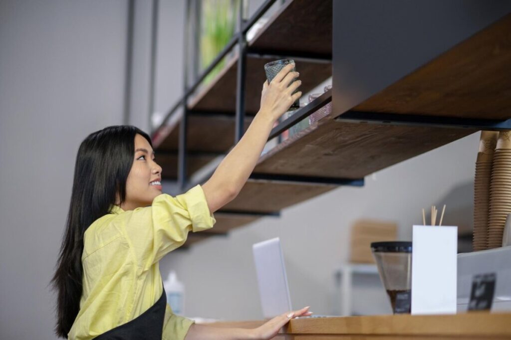 A barista in a yellow shirt reaches up to grab a glass from a wooden shelf in a modern café.