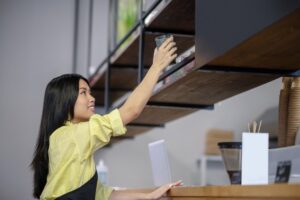 A barista in a yellow shirt reaches up to grab a glass from a wooden shelf in a modern café.